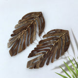 Pair of wooden leaf-shaped earrings on a white background with a green plant leaf.