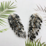 Pair of feather-shaped earrings on a white background with green palm leaves.
