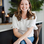 Woman sitting indoors wearing a white blouse with ruffled sleeves, smiling.