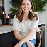 Woman sitting indoors wearing a white blouse with ruffled sleeves, smiling.