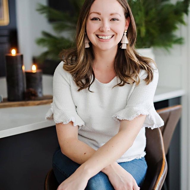 Woman sitting indoors wearing a white blouse with ruffled sleeves, smiling.