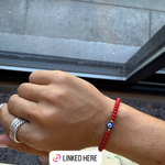 Hand wearing a red bracelet with a blue bead, silver ring, and silver chain, with text overlay on an Instagram post.