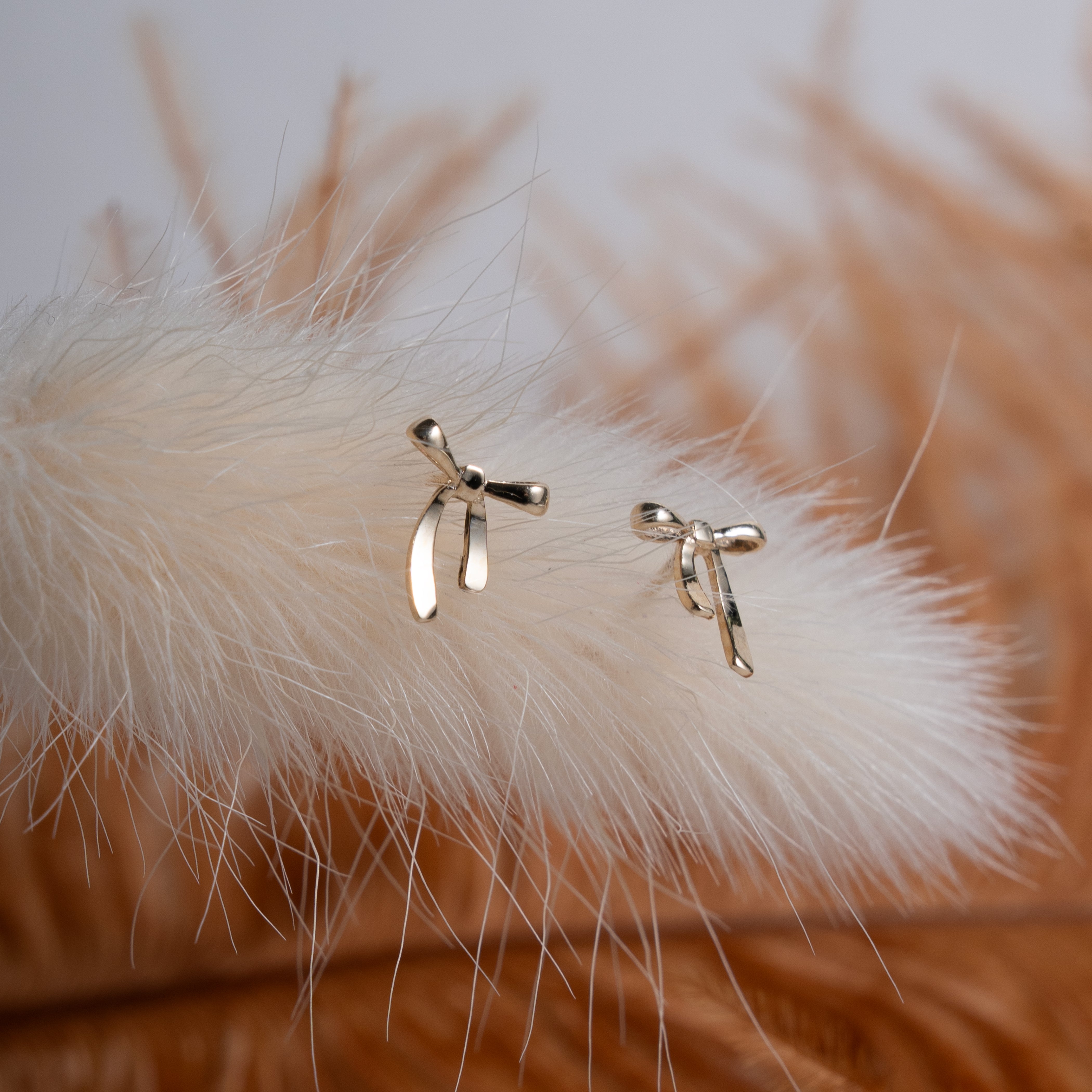 Gold bow-shaped earrings on a fluffy white surface with a blurred natural background